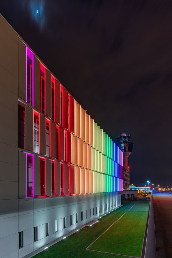 Long horizontal view of an airport terminal facade lit by a rainbow of vertical LED panels at night