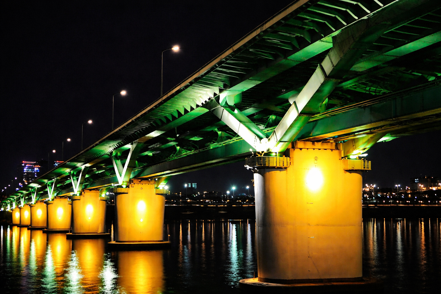 Modern bridge structure illuminated by green and yellow LED flood lights at night, showing uniform light distribution and reflections on the water.